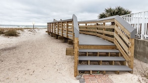 A wooden staircase leading to a beach area with a sandy path and a white fence in the background.