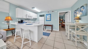 A modern kitchen with white cabinets, a microwave, and a sink. There is a dining area with a table and chairs, and a living room with a sofa and a lamp.
