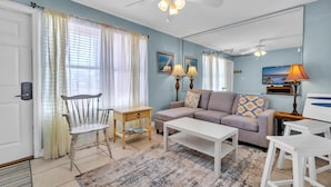 A living room with a white ceiling fan, a beige sofa, a white coffee table, and a wooden chair. There are two wall-mounted lamps and a television on the wall.