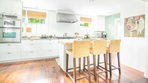 A modern kitchen with white cabinetry, a central island with bar stools, a stove, and a large window with blinds. - Spend the Night in an Historical, Luxury Downtown Home by Lucky Savannah (Savannah)