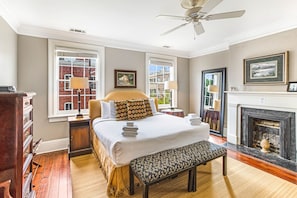 Quarto com cama grande, um banco, lareira e vista da cidade pelas janelas.
