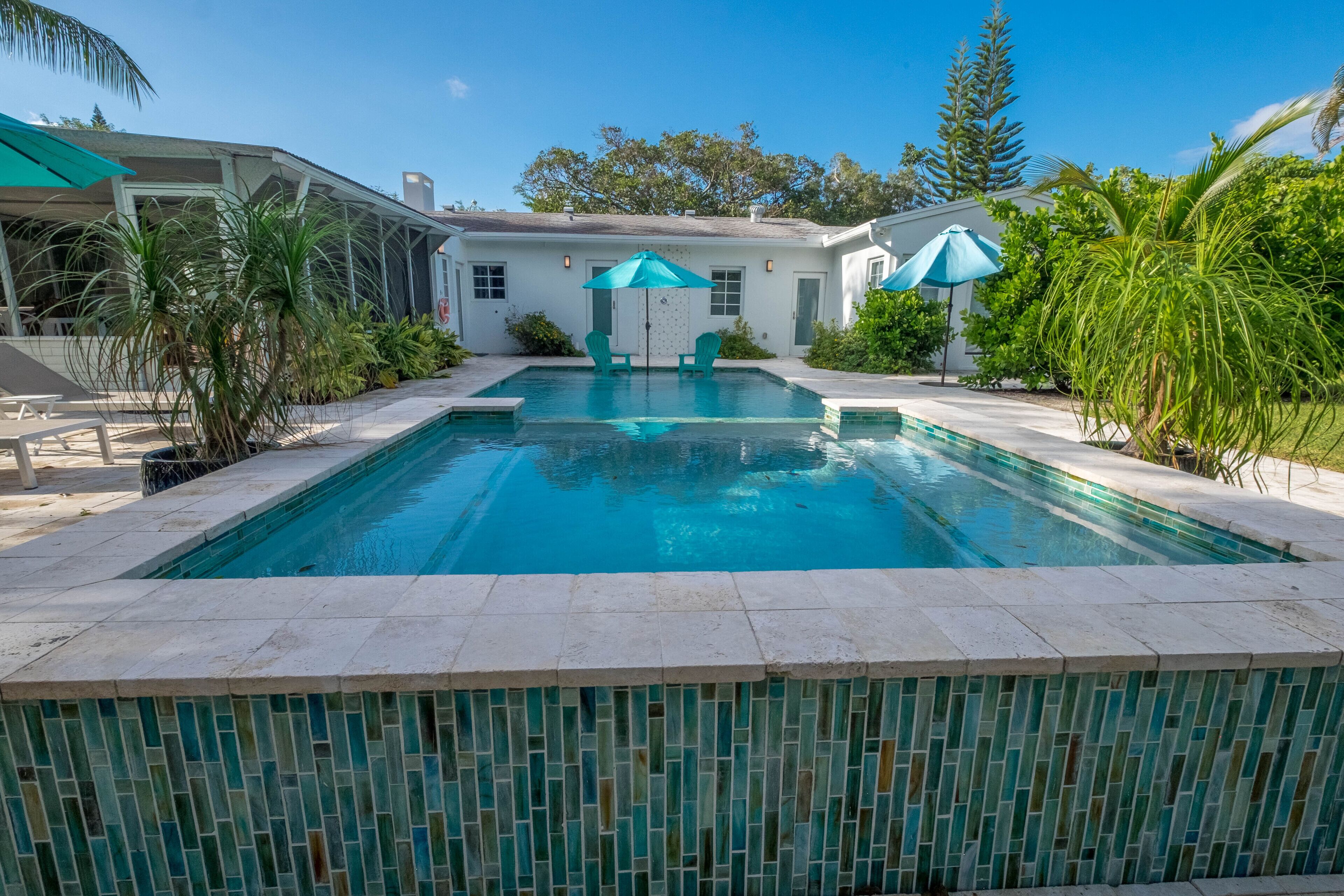 Une piscine bordée de carreaux, deux parasols verts et une maison en arrière-plan.