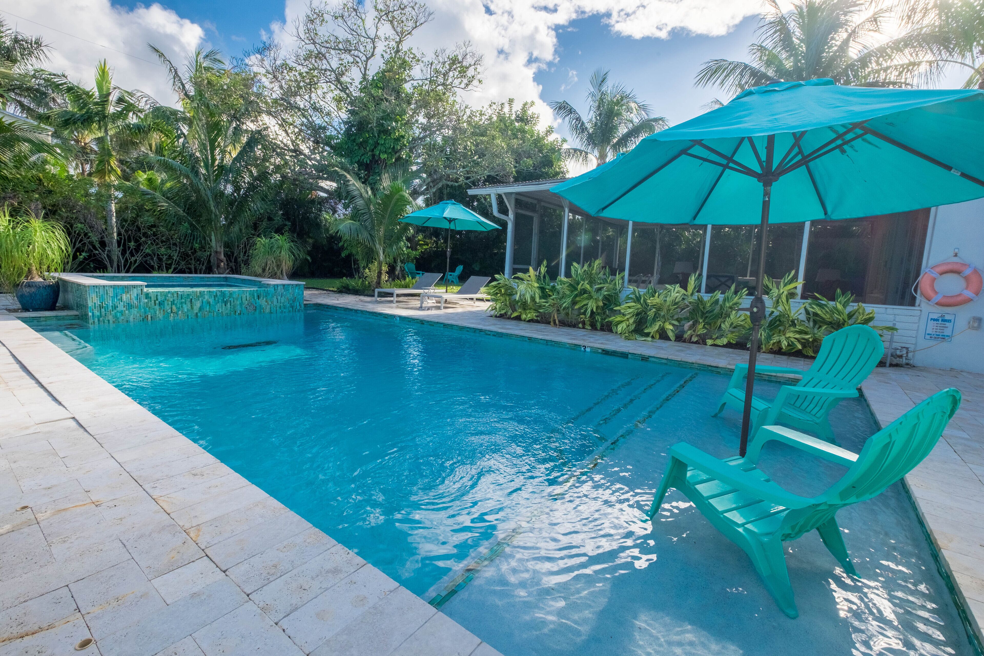Una piscina con agua azul cristalina, rodeada de plantas en macetas y dos sombrillas verdes.