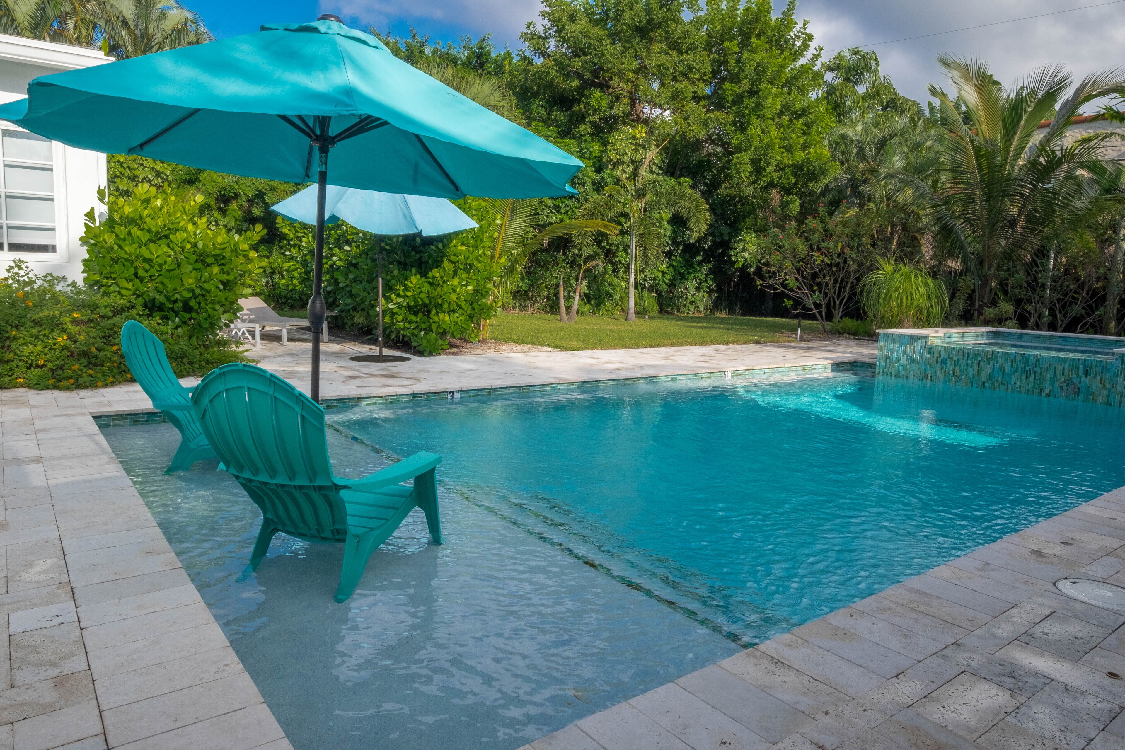 A swimming pool with a turquoise umbrella and chairs, surrounded by lush greenery.