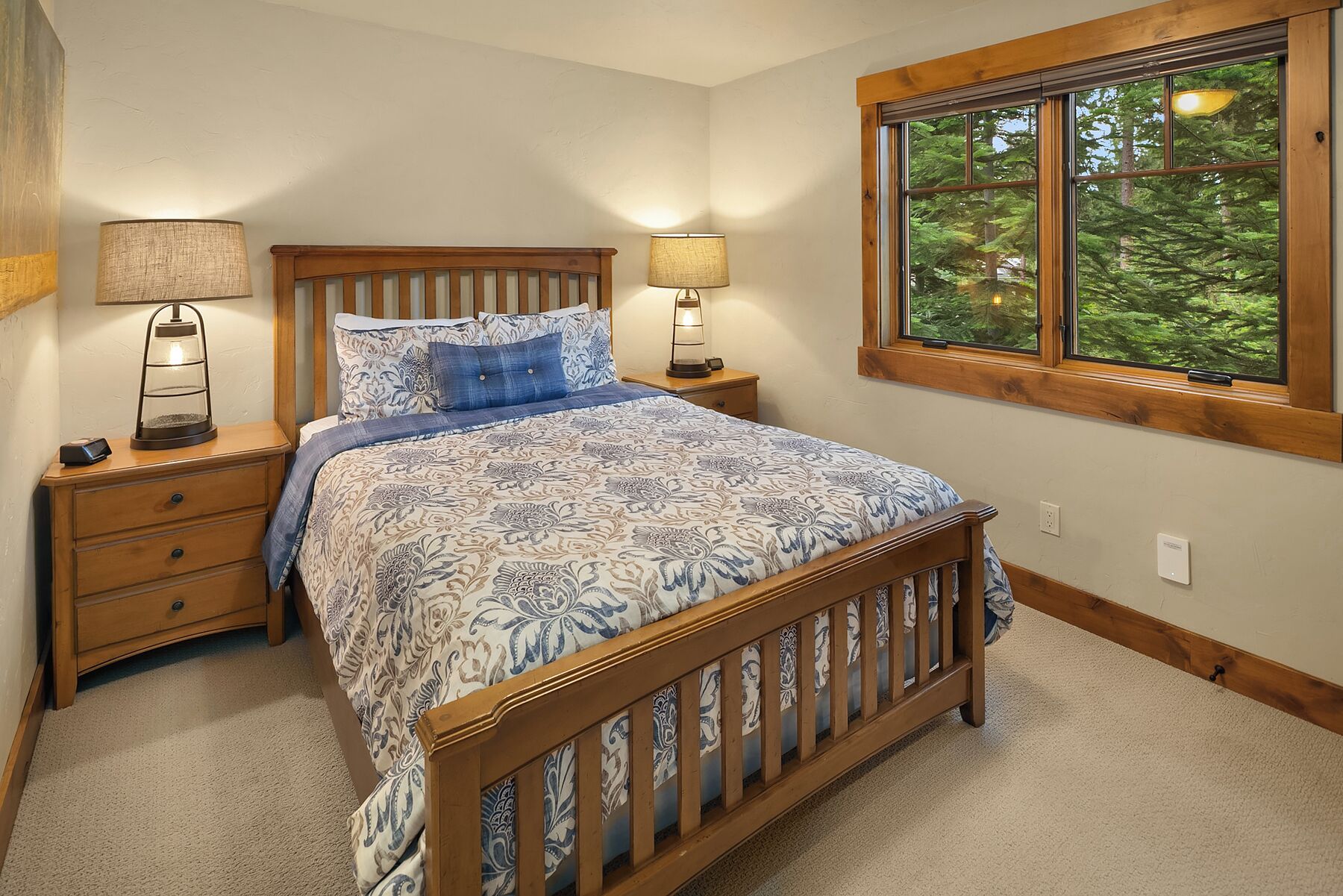 Un dormitorio con cama de madera, dos mesitas de noche con lámparas, una ventana con vista a árboles y suelo alfombrado.