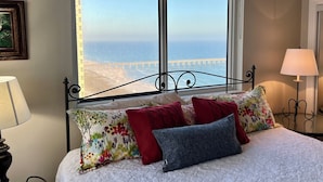 A bedroom with a large window overlooking a beach and pier, a bed with floral and solid-colored pillows, and a bedside lamp. - Wilson's Beach Dream: Gulf Front 5 Star 3BR 3Bath with Beach Service (Navarre)