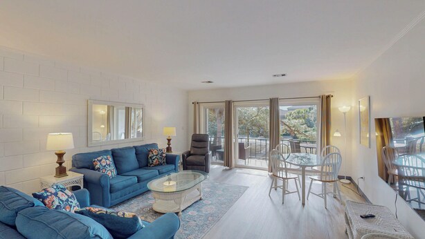 A modern living room with a blue sofa, a glass coffee table, and a dining area with a glass table and chairs. - Atlantic Breeze #5 2 bedrooms, 2 bathrooms (Jekyll Island)