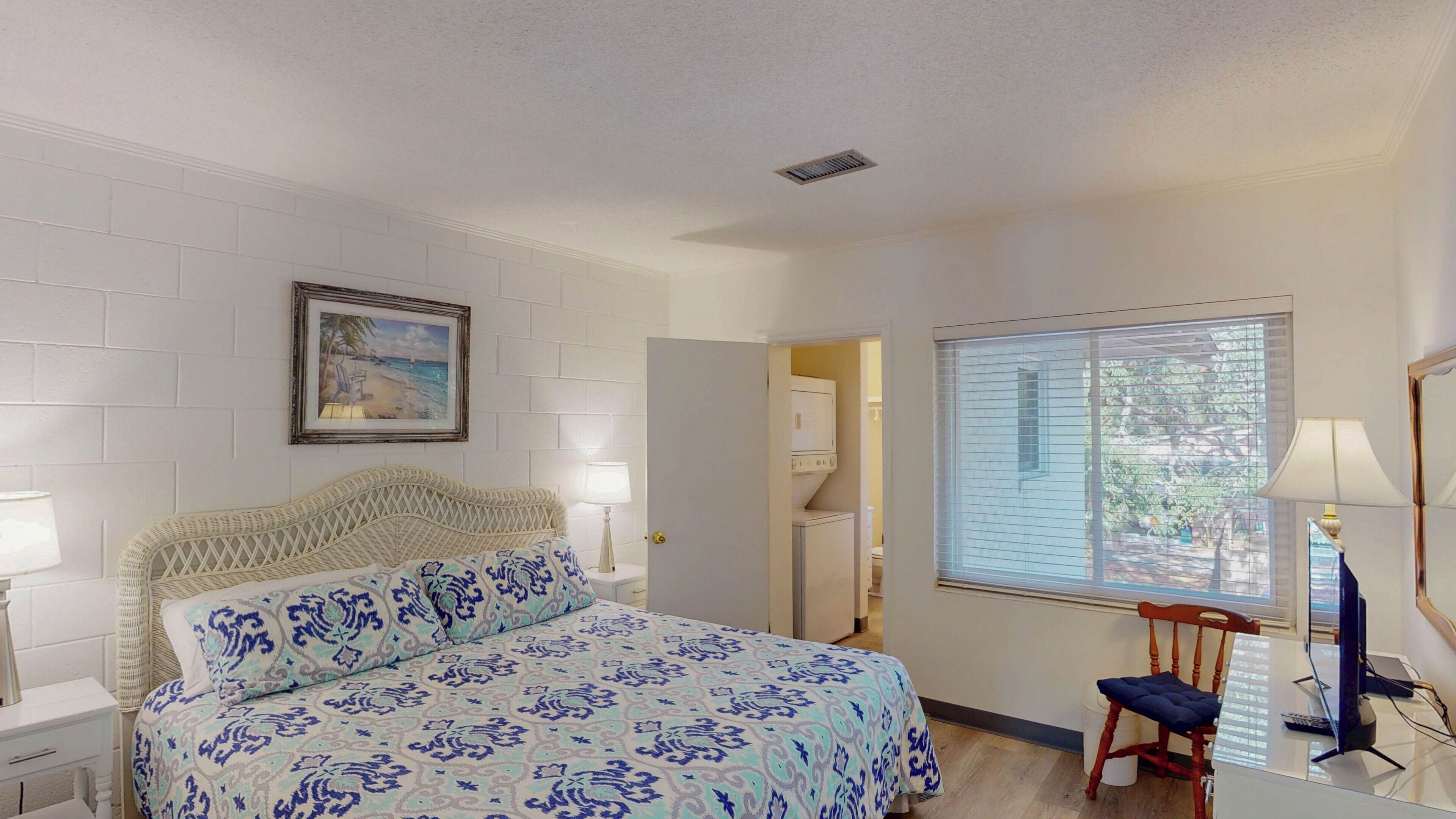 A bedroom with a bed, a nightstand, a lamp, a chair, a desk with a computer, a window with blinds, and a door leading to a bathroom.