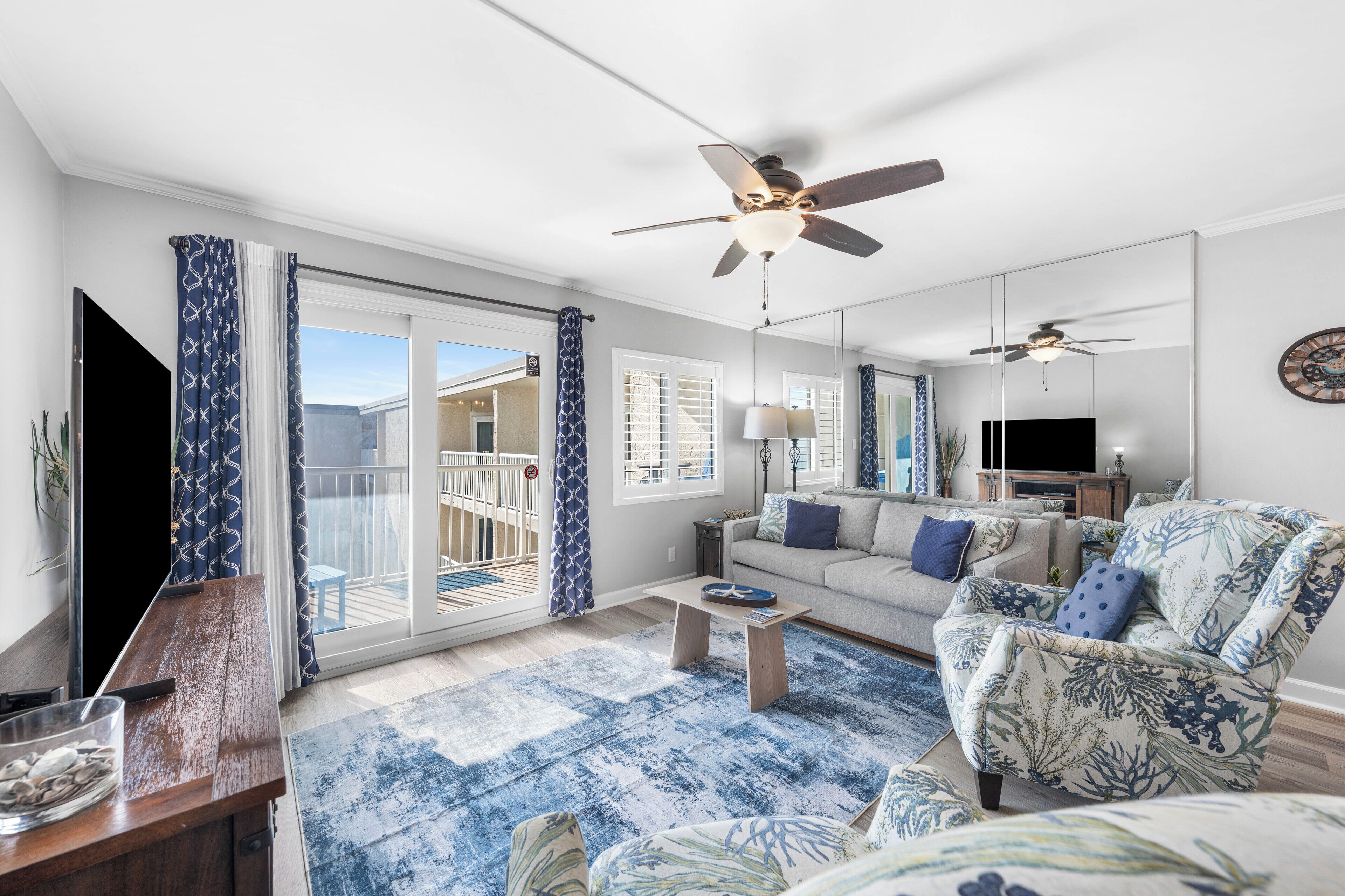 A spacious living room with a large sofa, a coffee table, and a ceiling fan. There is a sliding glass door leading to an outdoor area.