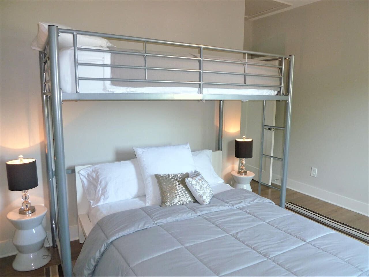 A bunk bed with a metal frame, white bedding, two bedside tables with lamps, and a plain wall.