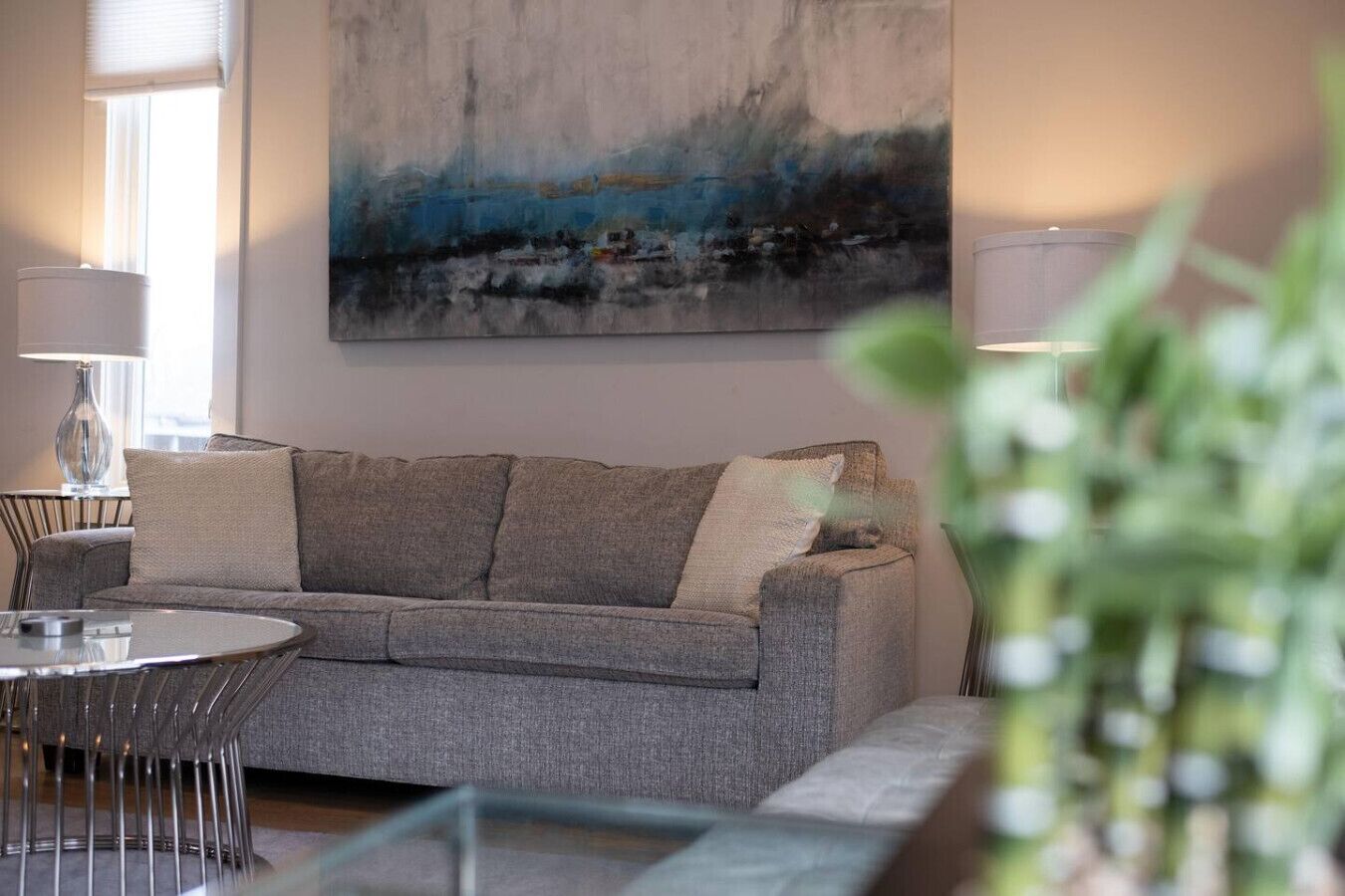 A modern living room with a grey sofa, a round glass coffee table, and a painting on the wall.
