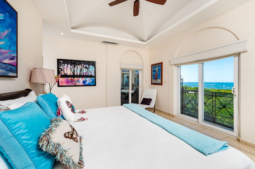 A bedroom with a large bed, a ceiling fan, a view of the ocean, and artwork on the walls.