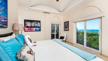 A bedroom with a large bed, a ceiling fan, a view of the ocean, and artwork on the walls.