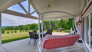 A covered patio with a table and chairs, a swing, and a view of a green landscape. - Rapalee Retreat - Wine and Lake Lovers Take Notice! (Himrod)