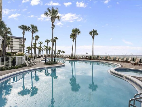 A swimming pool with palm trees and lounge chairs, surrounded by a building with a balcony.