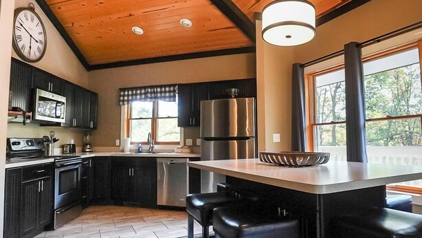 A modern kitchen with dark cabinetry, stainless steel appliances, a central island with bar stools, and a wooden ceiling.
