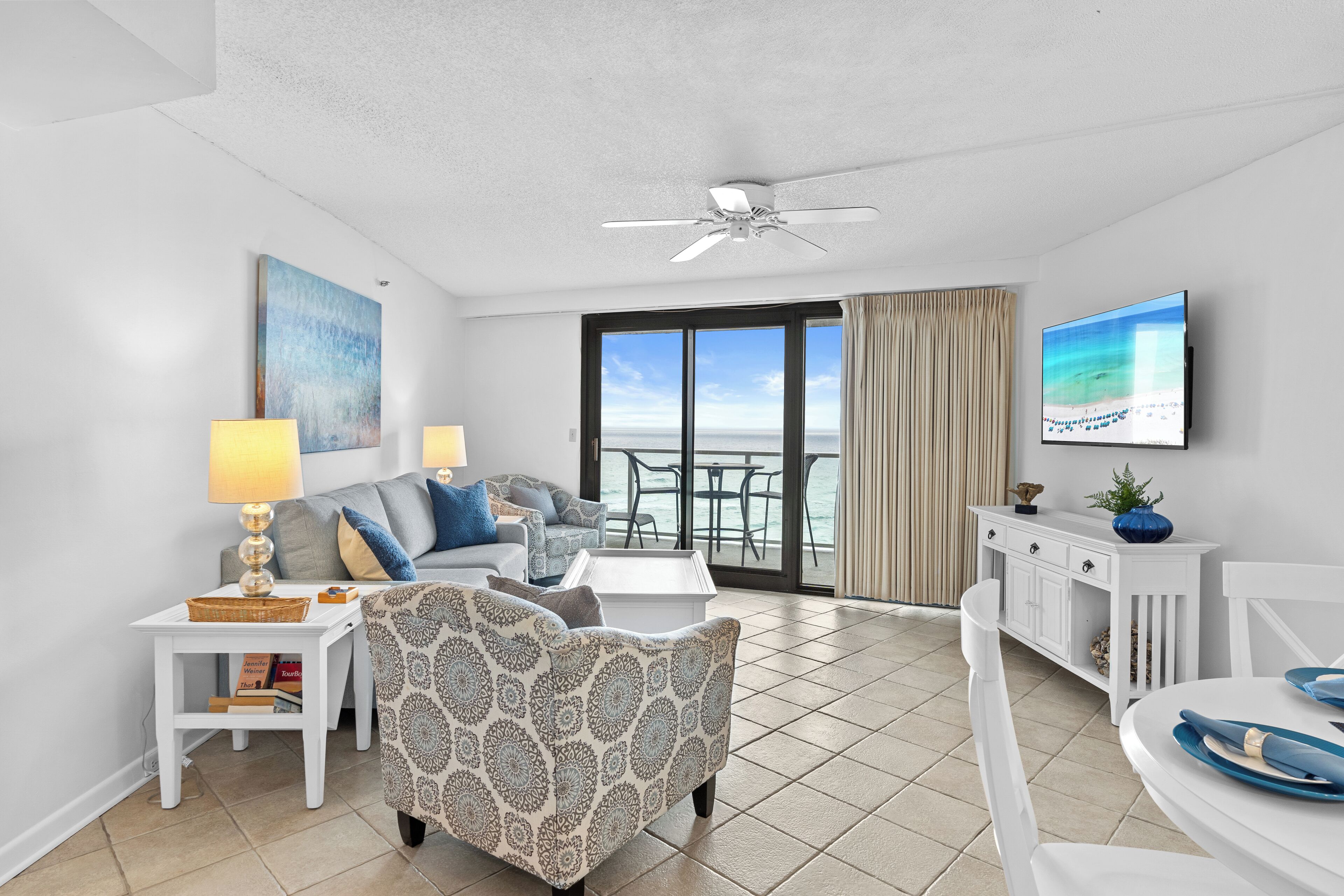 A modern living room with a sofa, armchair, and coffee table, a television, and a balcony with a view of the sea.