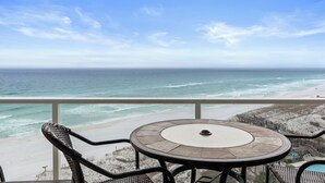 A balcony with a round table and two wicker chairs, overlooking a beach with waves and a clear sky. - Rate Reduced! Beachfront 2BR w/ Gulf Views, GOLF CART! Steps to Beach & Pool. (Destin)