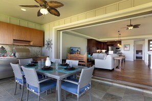 A spacious dining area with a high ceiling fan, a kitchen with wooden cabinets, and a living room with a sofa and a dining table set for a meal. - Savor The Best - Beautifully decorated with Golf Course And Maui Views (Kamuela)