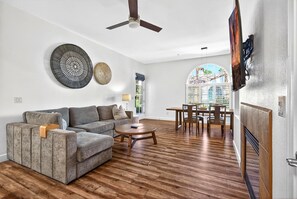 A living room with a sectional sofa, a round coffee table, a dining table with chairs, a ceiling fan, and a large window with a view.