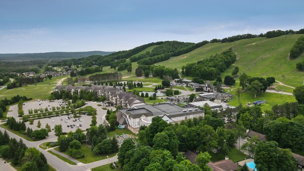 A large, historic building complex with multiple wings and a central courtyard, surrounded by lush green hills and a parking area.