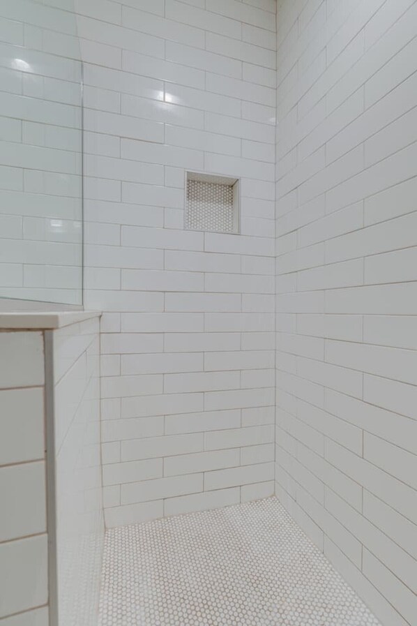 Une salle de bain avec des carreaux de céramique blancs rectangulaires aux murs et au sol, une petite fenêtre et un espace de douche carrelé.