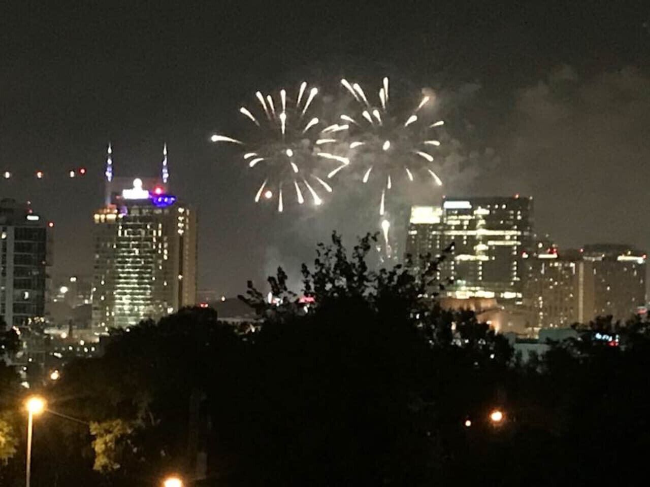 Une vue nocturne d’une ville, avec des feux d’artifice dans le ciel, des bâtiments illuminés et des arbres au premier plan.