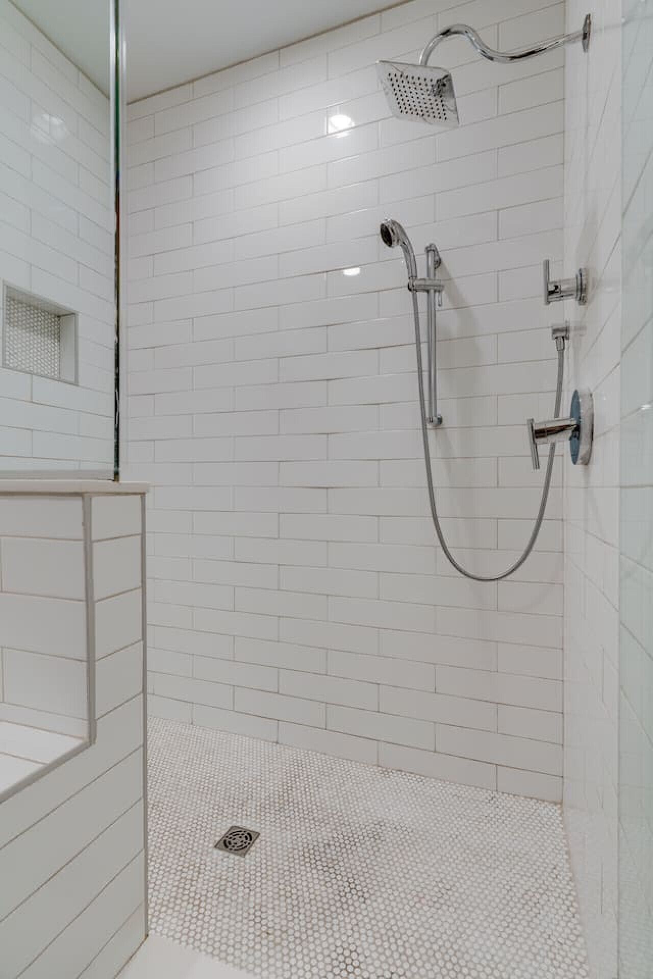 Un área de ducha moderna con paredes y suelo de azulejos blancos, cabina de ducha de vidrio y plato de ducha metálico.