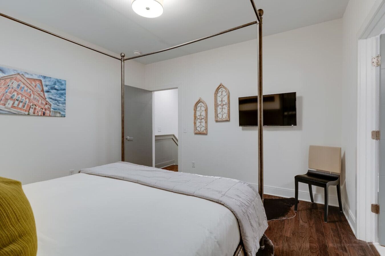 A bedroom with a four-poster bed, a television mounted on the wall, and a painting of a building on the left wall.