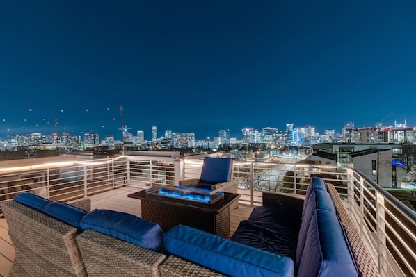 A rooftop terrace with a city skyline view, outdoor seating with cushions, and a table with a built-in heater. - The New Nashview: 4BR Gulch | Rooftop Fire Pit (Nashville)