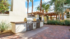 Una cocina al aire libre con una parrilla y armarios de almacenamiento, rodeada de palmeras y una pérgola.