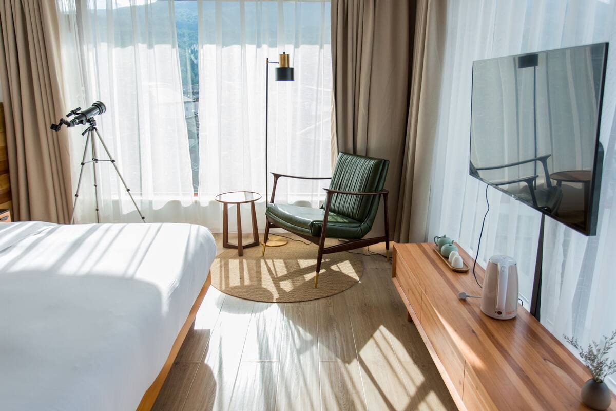 Habitación de hotel con una cama, una silla verde, un televisor, un telescopio y vistas a la ciudad a través de amplios ventanales.