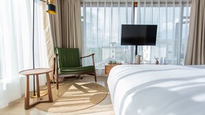 A hotel room with a large bed, a green armchair, a small round table, a TV, and a view of buildings through sheer curtains. - Wangshan Travelling With Hotel (Lijiang)