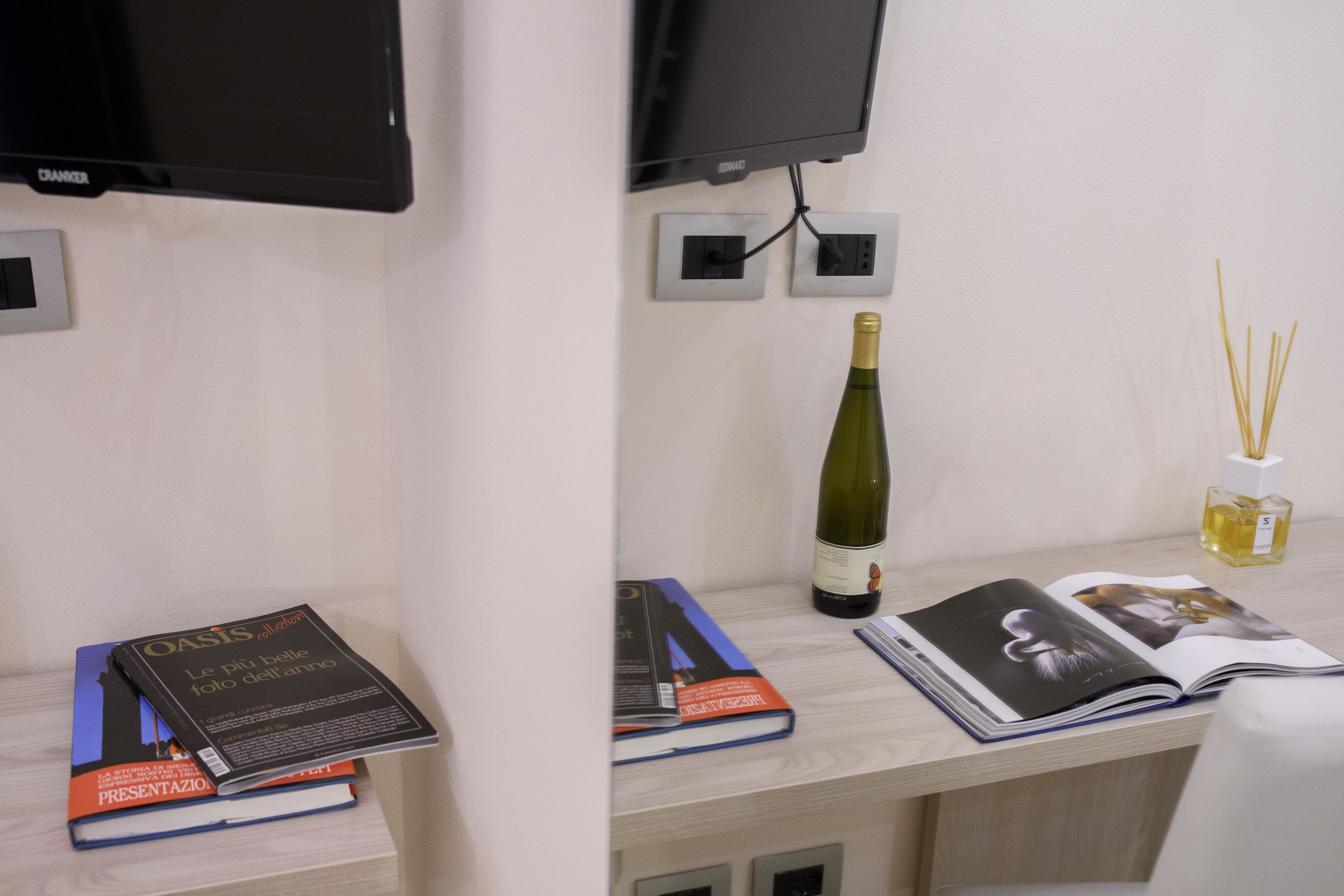 Habitación de hotel con un escritorio de madera, un televisor montado en la pared, una botella de vino, una revista y algunos libros.