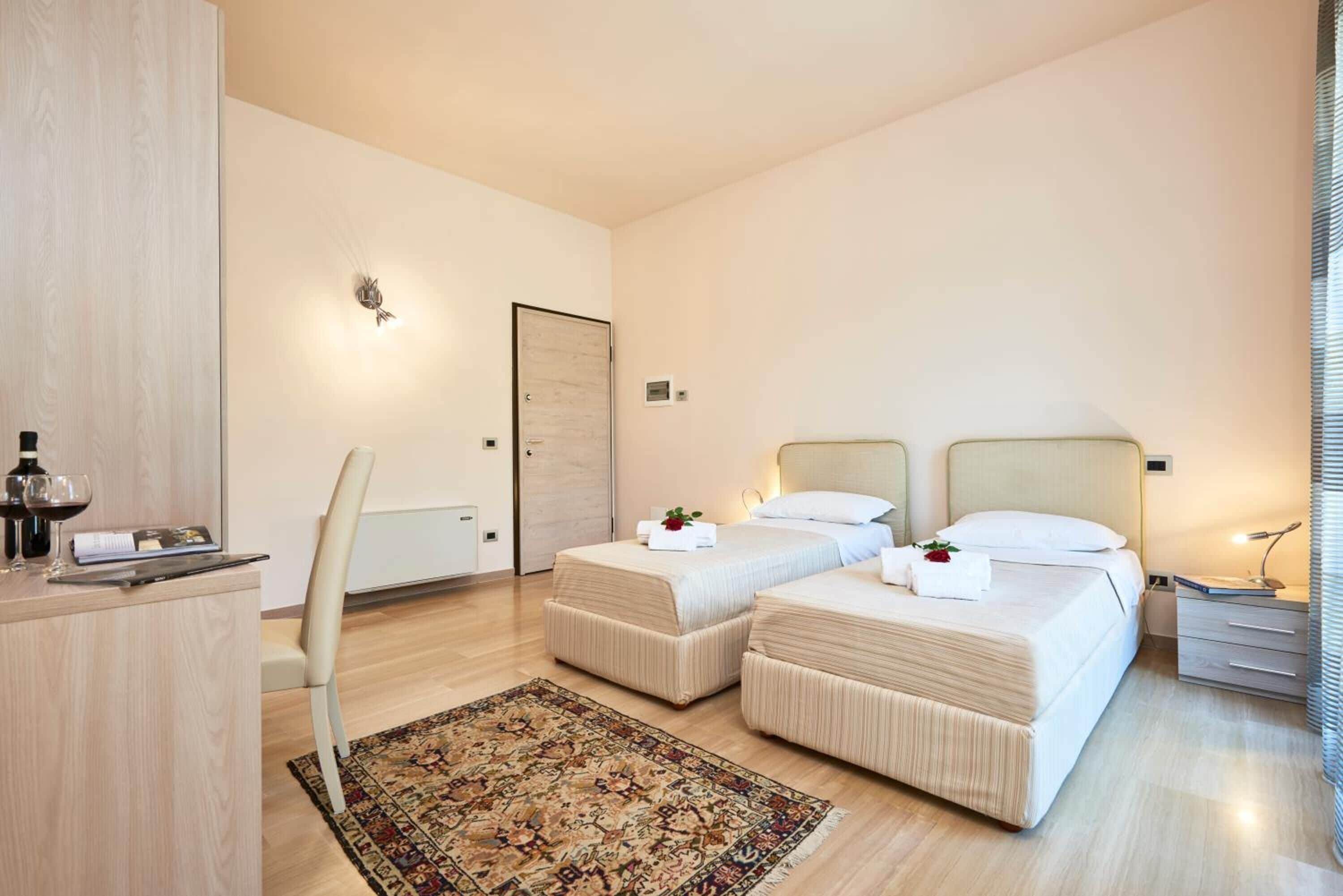 Habitación de hotel con dos camas, un escritorio, una silla, una mesita con lámpara y una alfombra decorativa.
