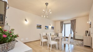 A modern dining area with a wooden table and white chairs, a chandelier, and a potted plant with pink flowers. - Domus il Palio B&B (Siena)