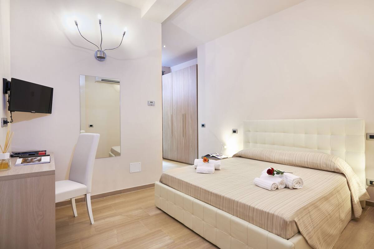 A modern hotel room with a large bed, a desk with a flat-screen TV, a chair, and a mirror.