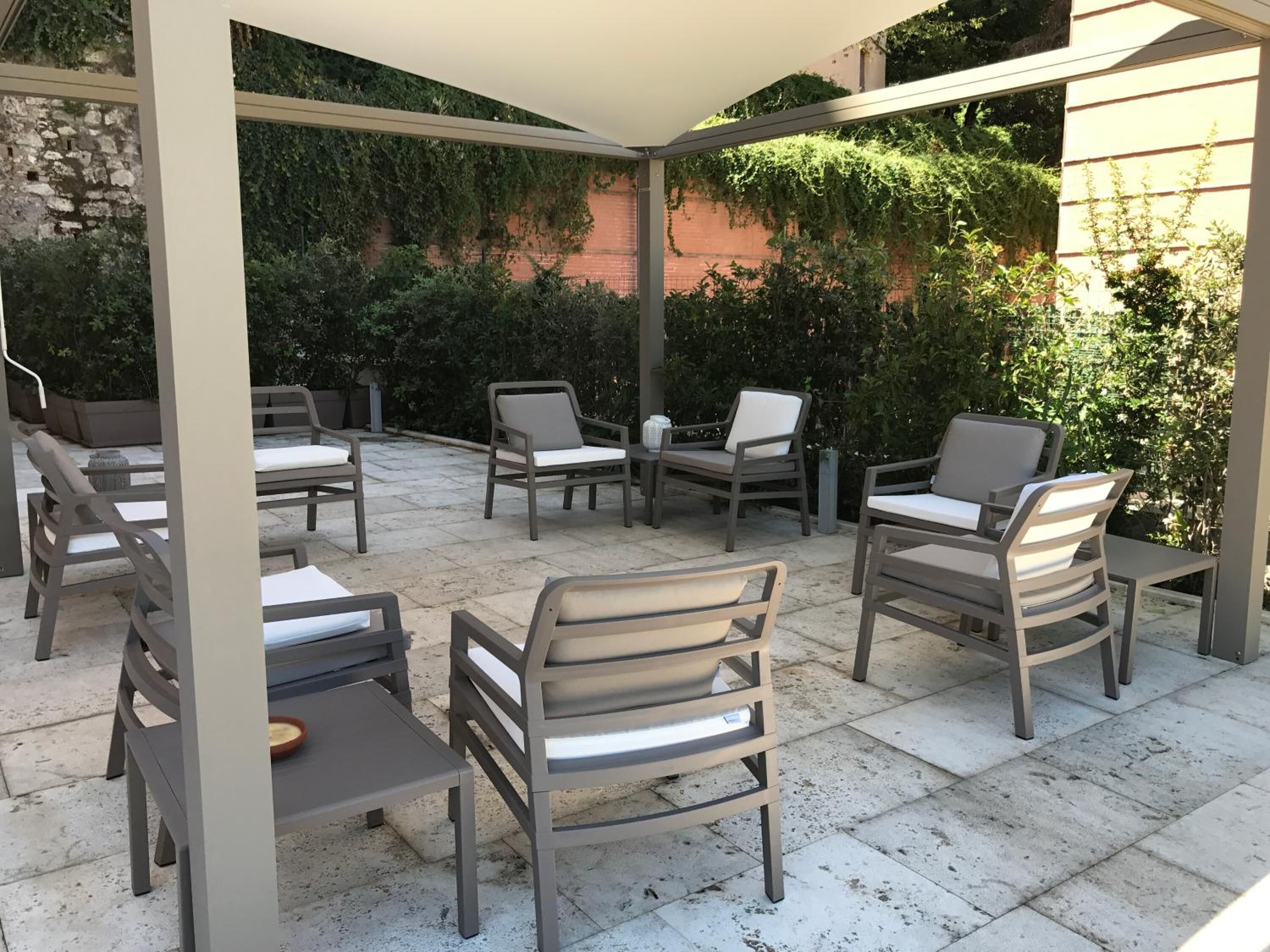 Patio esterno con diverse sedie e tavoli, immerso nel verde e circondato da un muro in mattoni.
