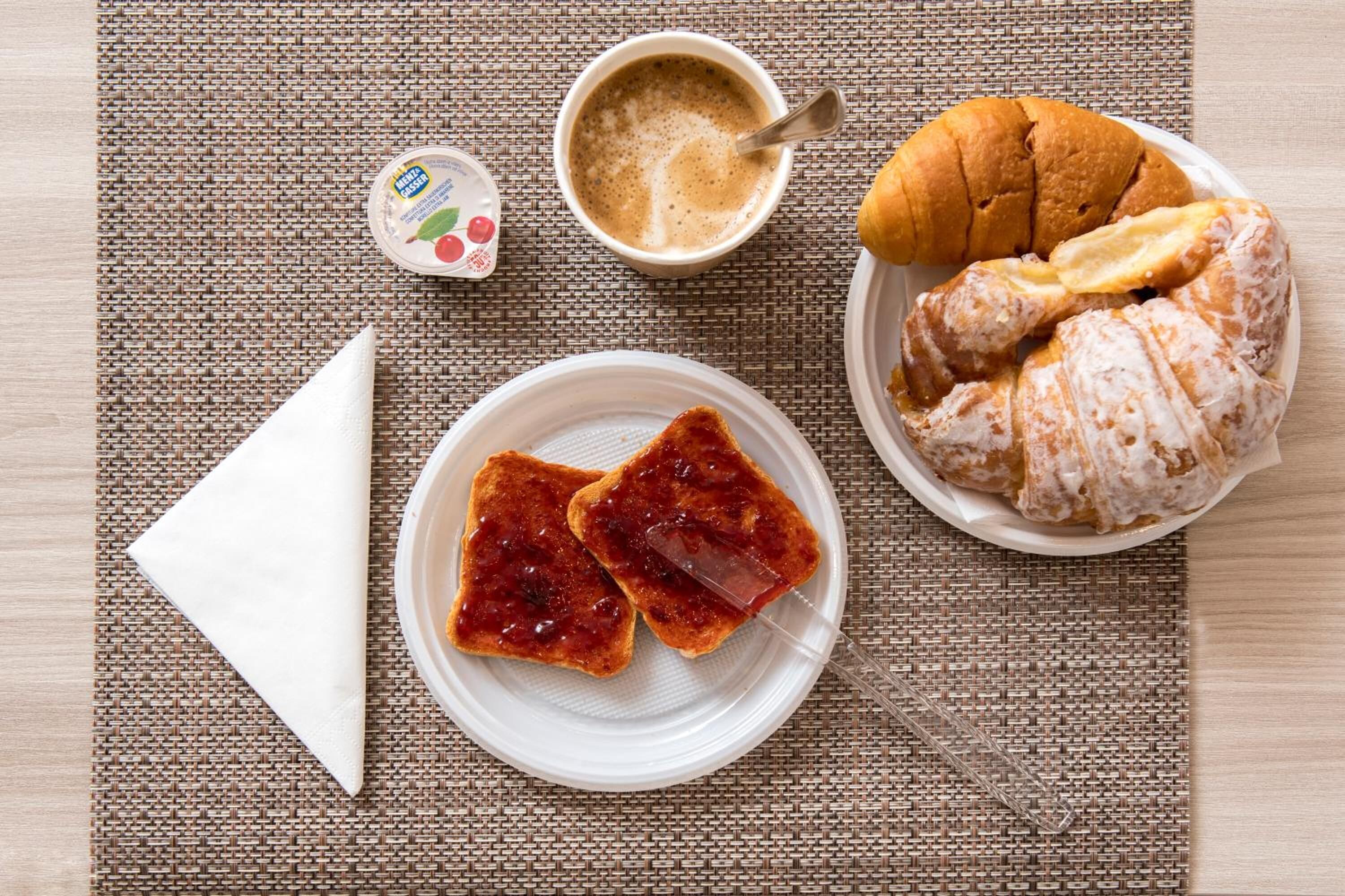 A breakfast setting with toast, jam, a croissant, and a cup of coffee on a woven placemat.