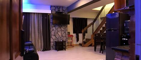 Amplia sala de estar con pared de acento de piedra, televisor de pantalla plana, chimenea y escalera que conduce a un nivel superior.