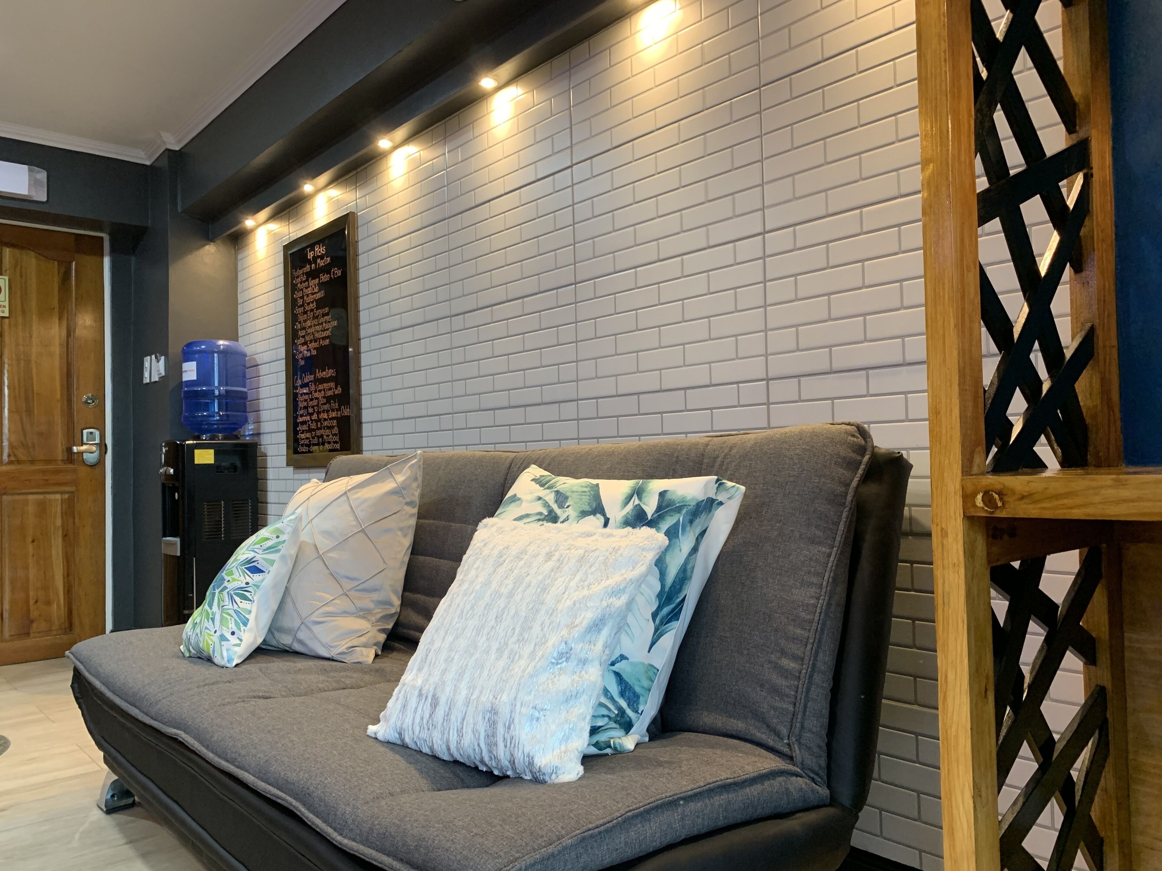 A modern living room with a grey sofa, two patterned cushions, a water dispenser, and a brick-patterned wall.