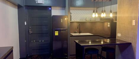 Cocina compacta con refrigerador negro, lavamanos y barra con taburetes.