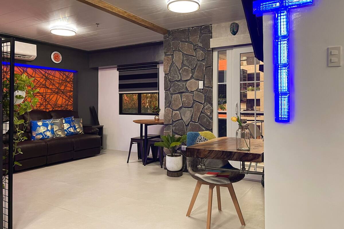Una sala moderna con un muro de acento de piedra, una zona de comedor con una mesa de madera y una cruz iluminada en azul.
