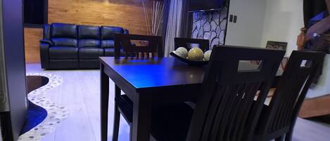 A modern living room with a dark wooden dining table and chairs, a black leather sofa, and a flat-screen TV mounted on the wall.