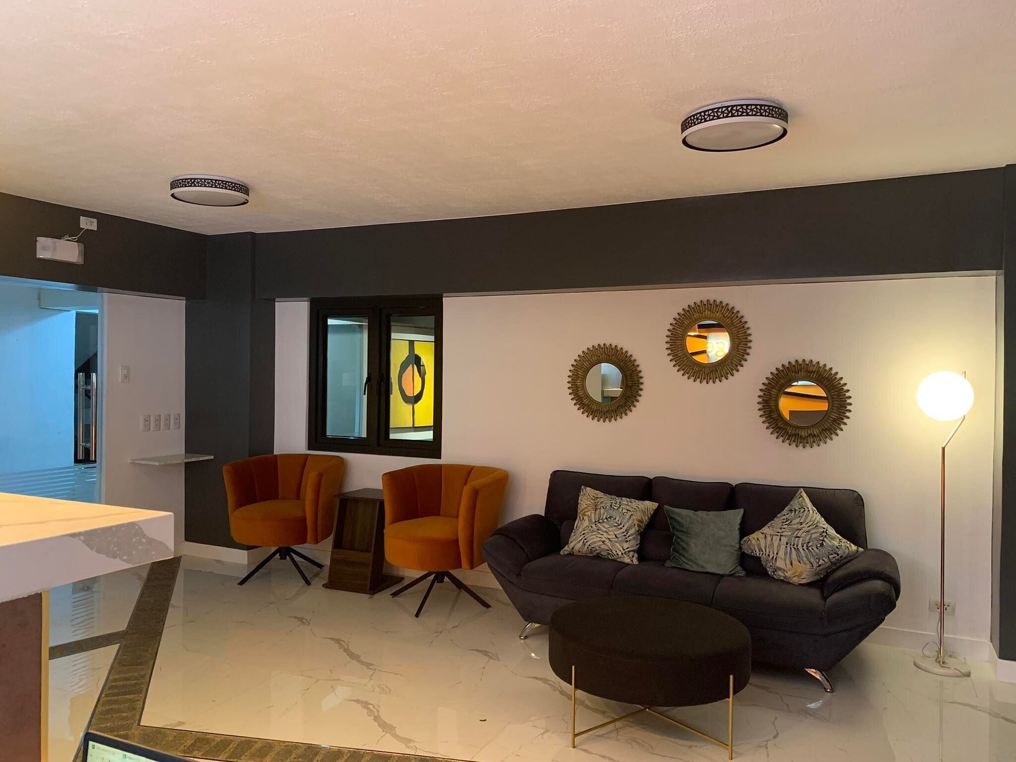 Un salon moderne avec un canapé de couleur foncée, deux fauteuils orange, un petit pouf rond et une lampe sur pied.