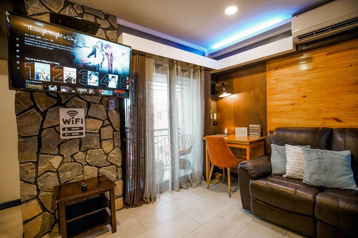 Una sala de estar acogedora con una pared de piedra, un televisor de pantalla plana mostrando un programa de viajes, una mesa de comedor de madera y un sofá de cuero.