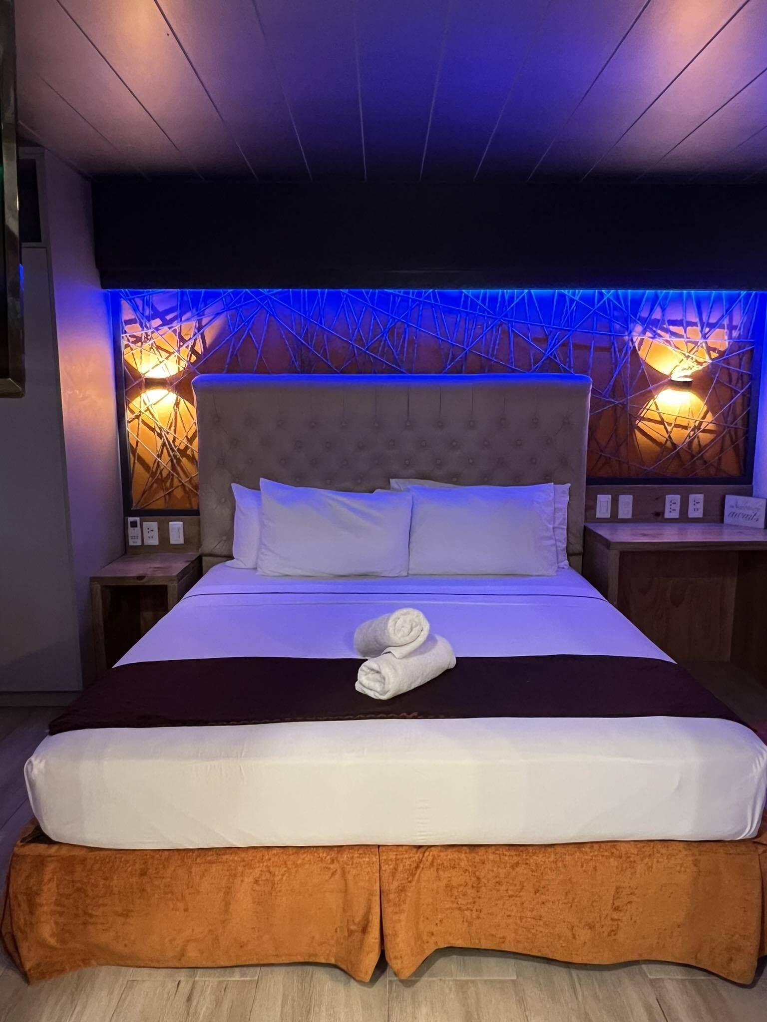 A neatly made bed with white linens and a brown skirt, a headboard with a geometric pattern, and two wall-mounted lamps with warm lighting.