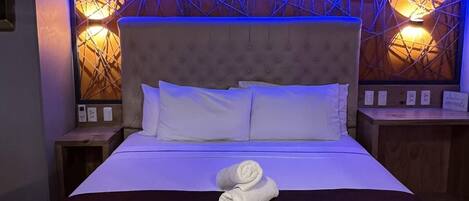 A neatly made bed with white linens and a brown skirt, a headboard with a geometric pattern, and two wall-mounted lamps with warm lighting.