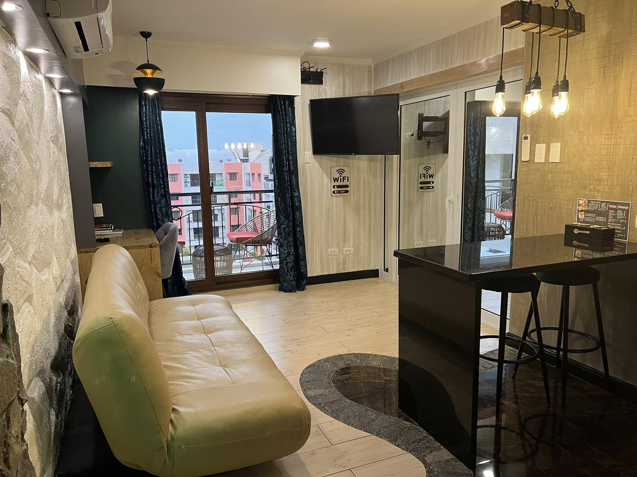 A modern living room with a sofa, a dining area with a bar, and a view of the outside balcony.