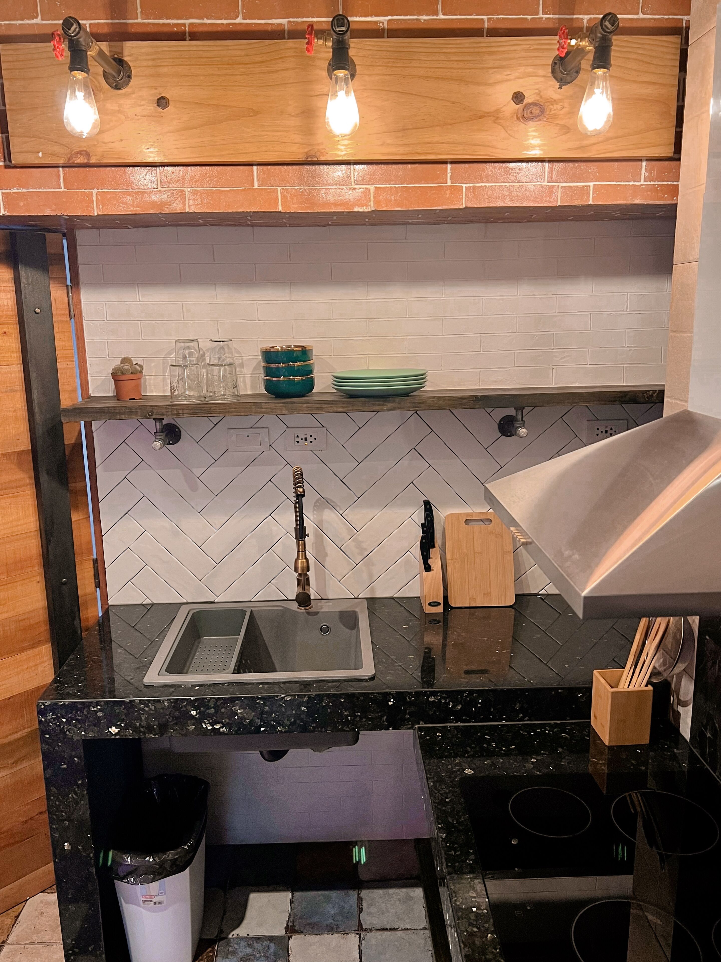 A kitchen with a black granite countertop, a stainless steel sink, a wooden shelf with glassware, and a modern stove.